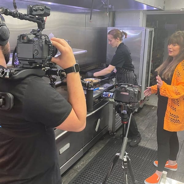 Filming in a commercial kitchen with a chef cooking on a griddle and a woman speaking.