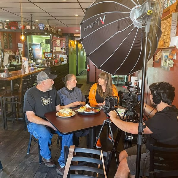 Three people at a restaurant table being filmed by a cameraman with a large softbox light.