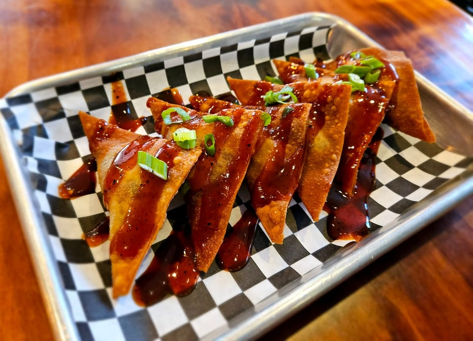 Crispy wontons drizzled with sweet and savory sauce, garnished with green onions on a checkered paper-lined tray.