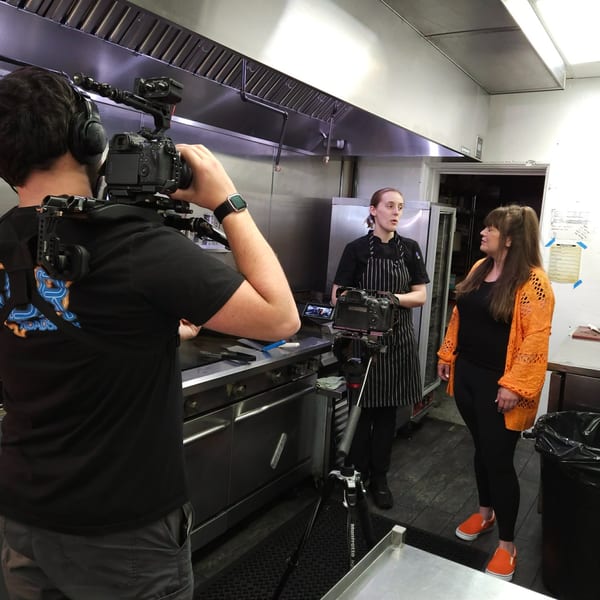A videographer films a chef and another woman talking in a commercial kitchen.