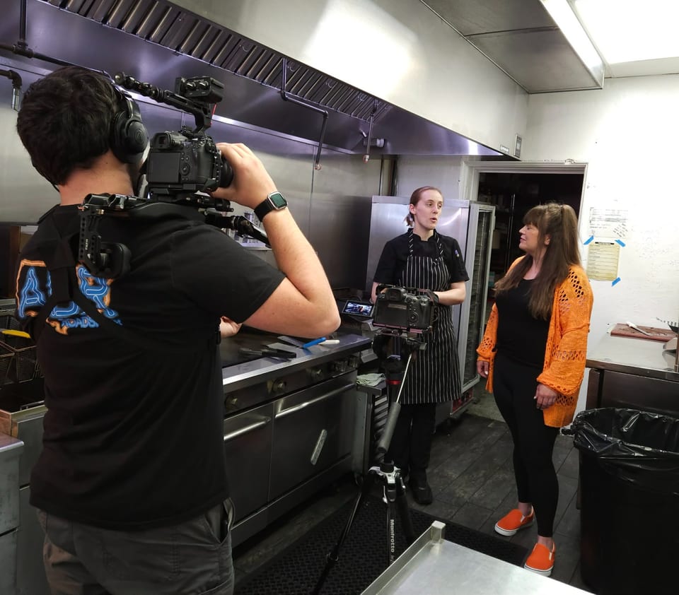 A videographer films a chef and another woman talking in a commercial kitchen.